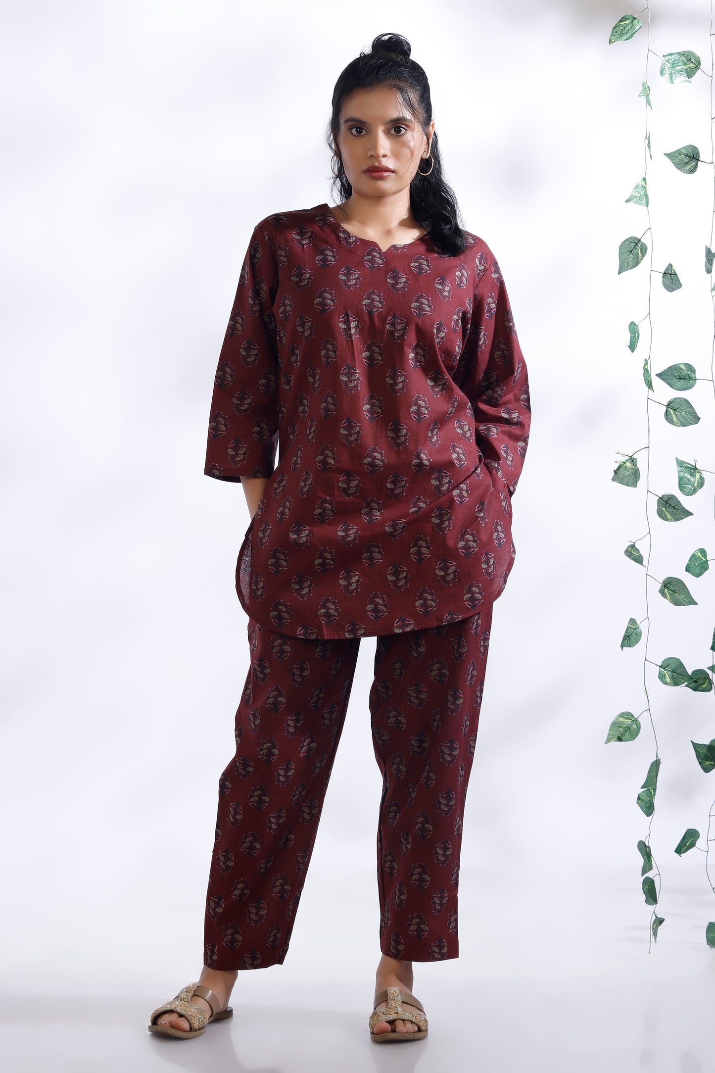 Woman wearing a maroon patterned outfit standing against a white background with a plant on the right side.