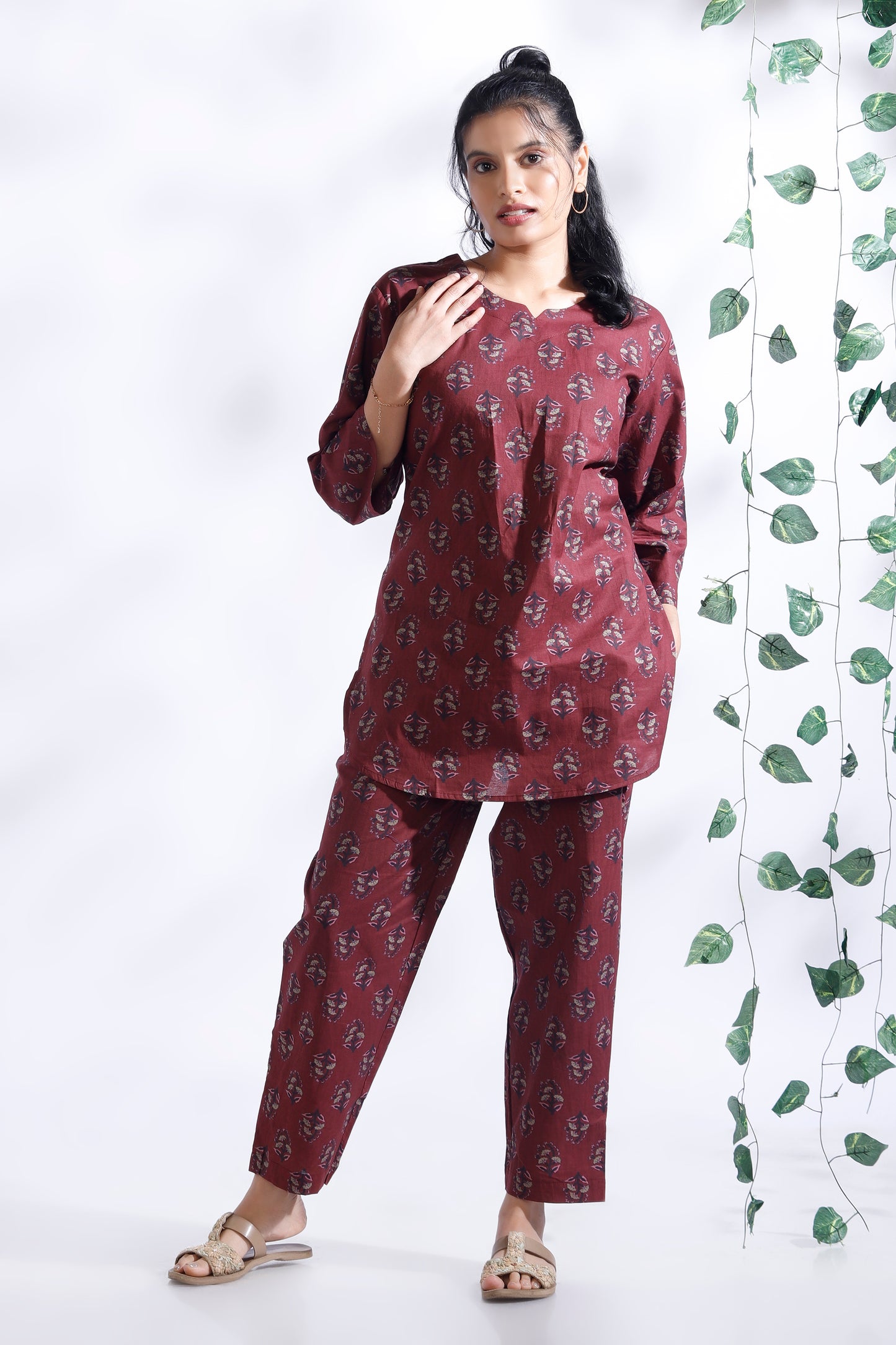Woman wearing a maroon patterned outfit standing next to a decorative plant on a white background