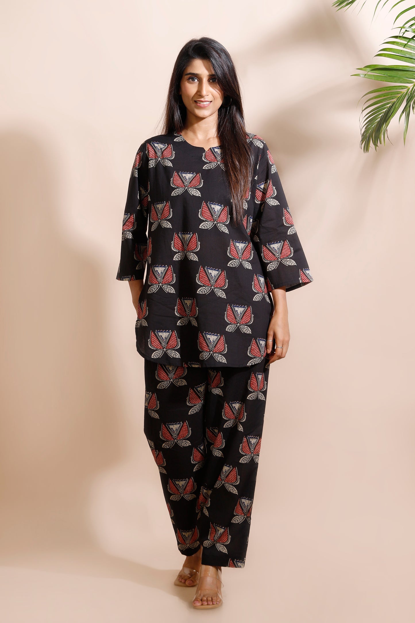 Woman wearing a black patterned outfit against a beige wall with a plant in the corner.
