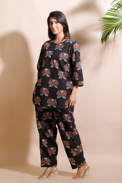 Woman wearing a black patterned outfit standing against a beige wall with a plant in the corner.