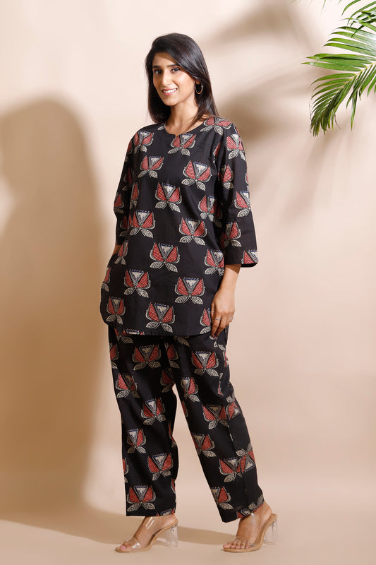 Woman wearing a black patterned outfit standing against a beige wall with a plant in the corner.