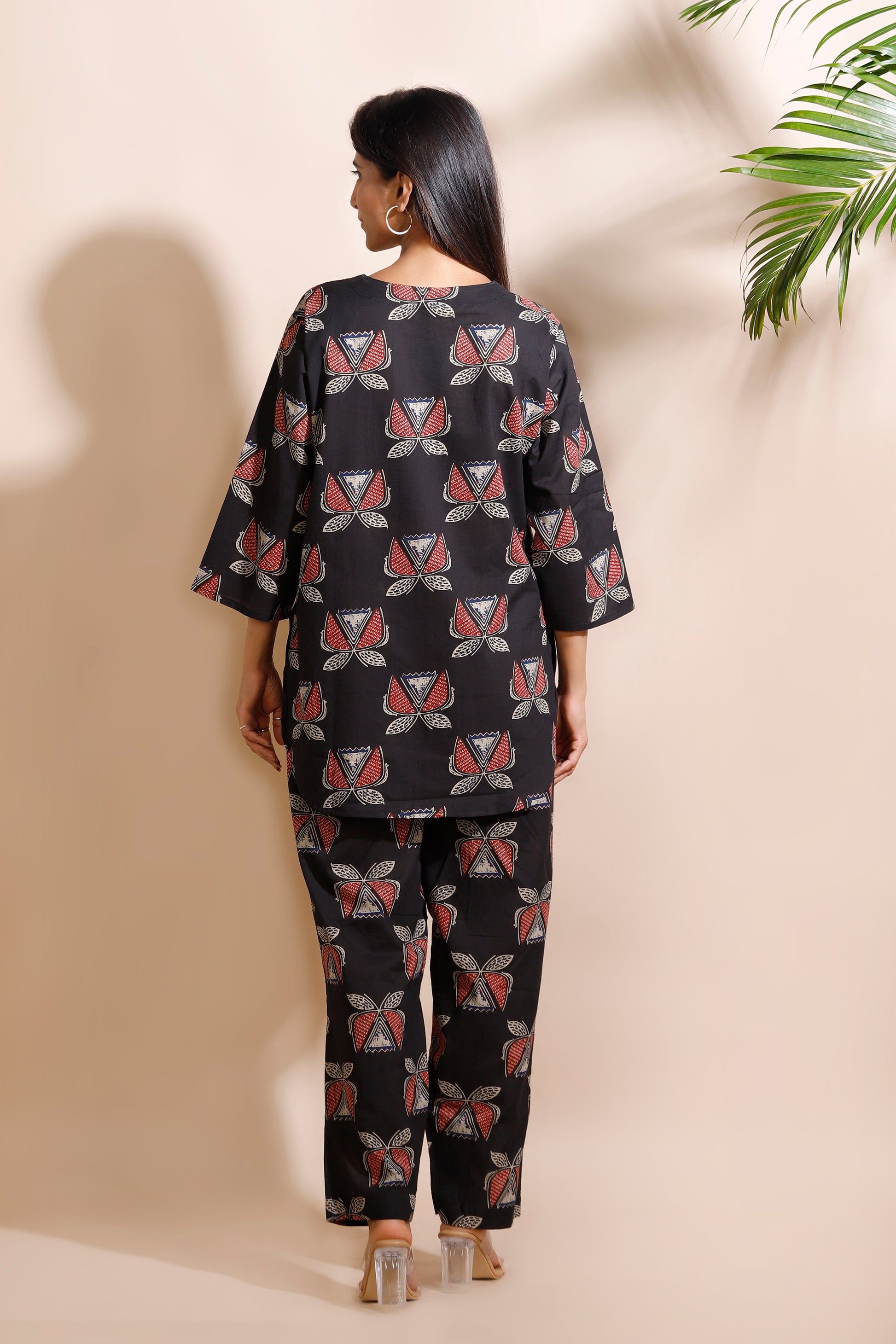 Woman wearing a black patterned outfit against a beige wall with a plant in the corner.