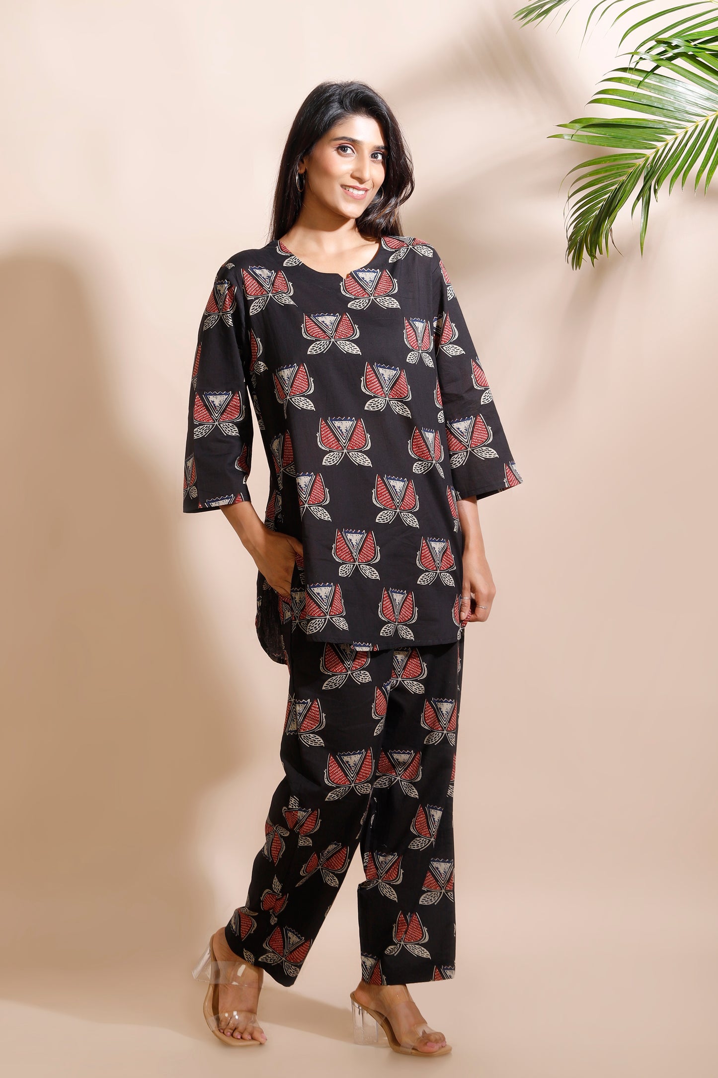Woman wearing a black patterned outfit against a beige wall with a plant in the corner.