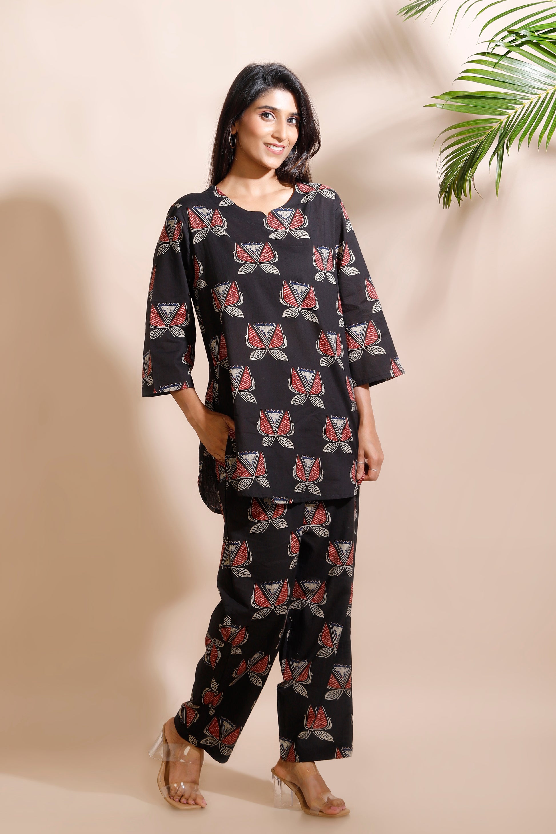 Woman wearing a black patterned outfit against a beige wall with a plant in the corner.