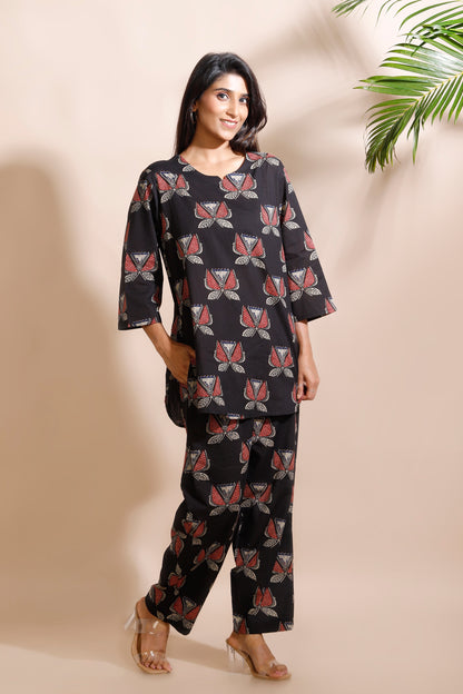 Woman wearing a black patterned outfit against a beige wall with a plant in the corner.