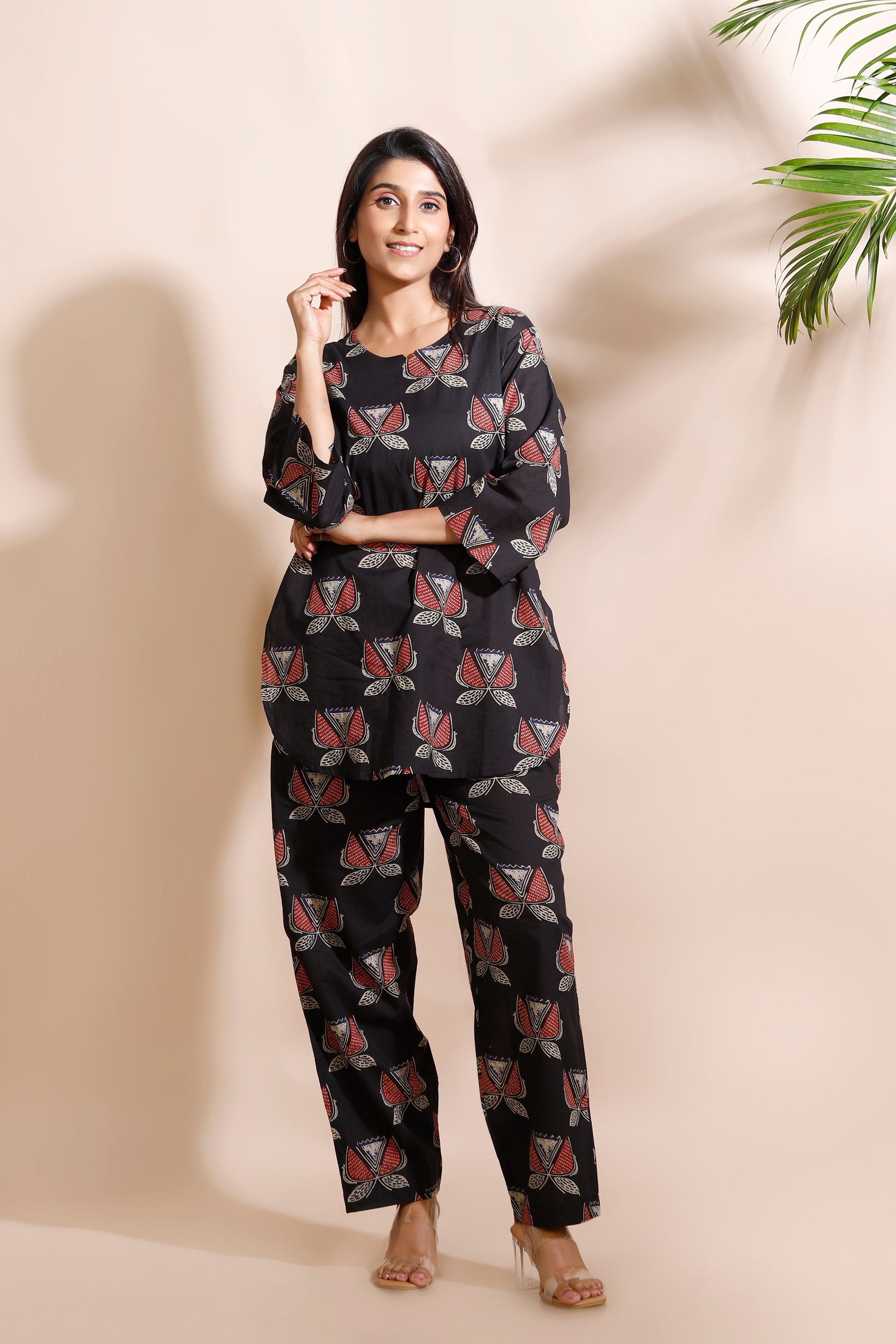 Woman wearing a black patterned outfit against a beige wall with a plant in the corner.