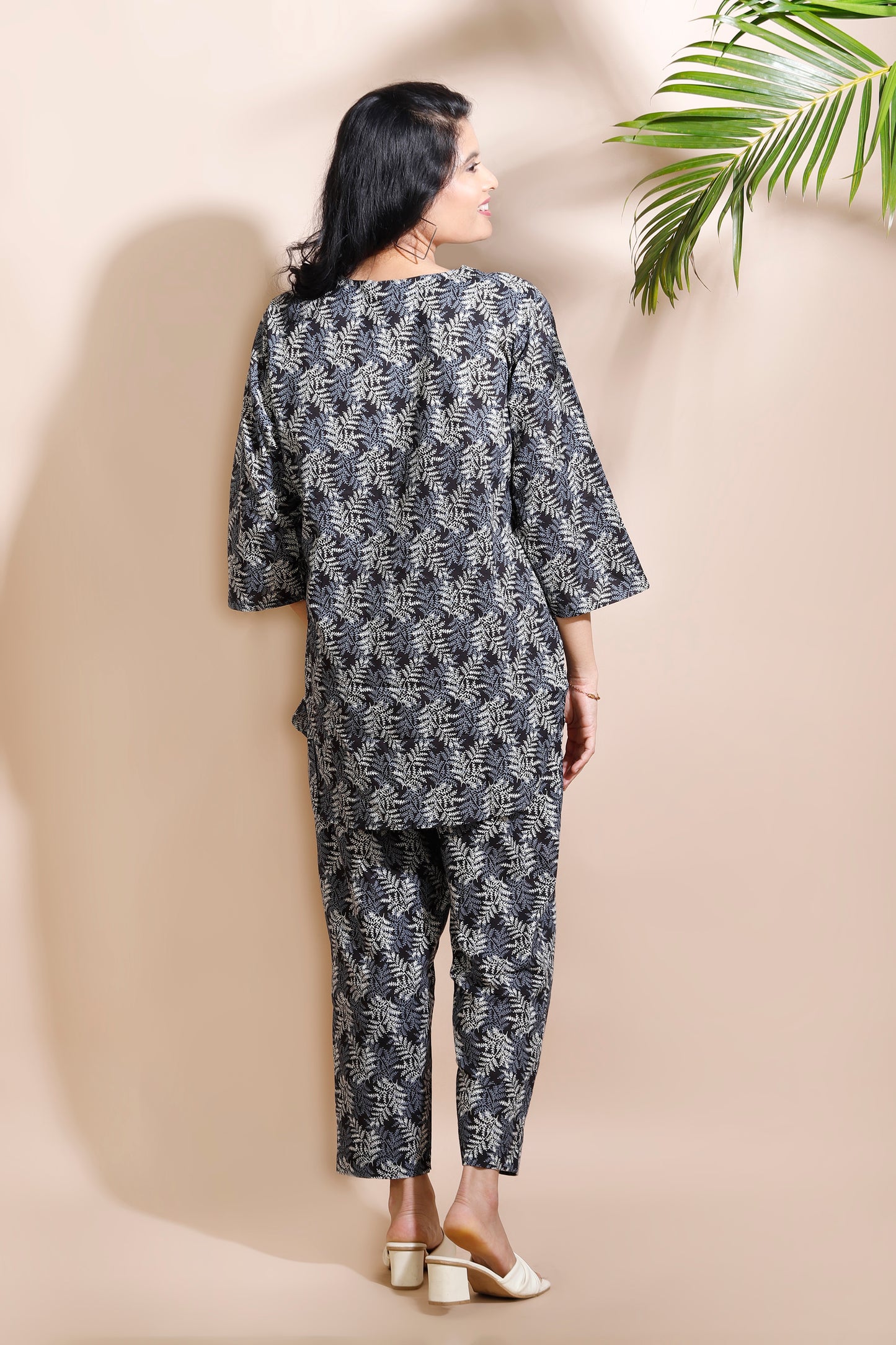 Woman wearing a black and white patterned outfit against a beige wall with a green leafy plant.