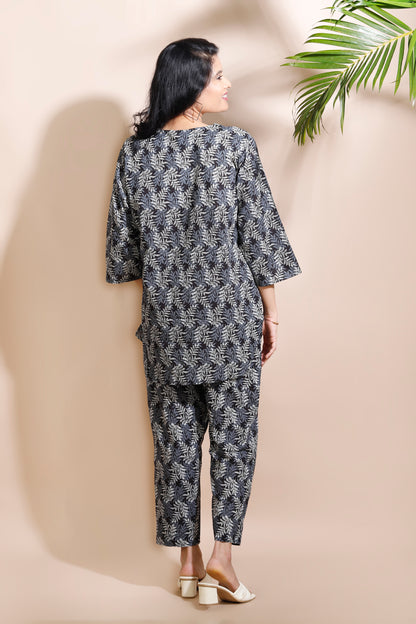 Woman wearing a black and white patterned outfit against a beige wall with a green leafy plant.