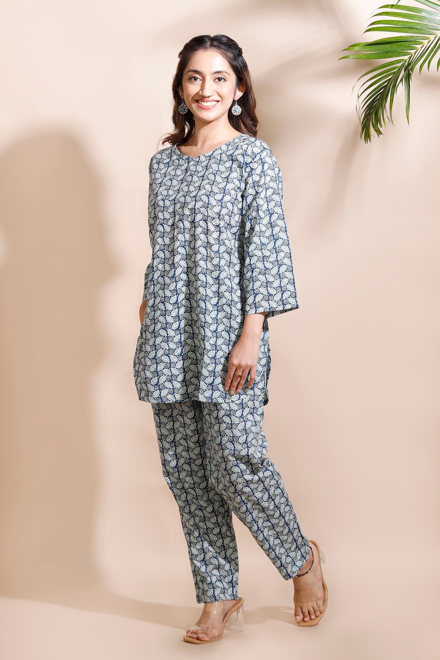 Woman wearing a blue patterned outfit standing against a beige background with a plant.