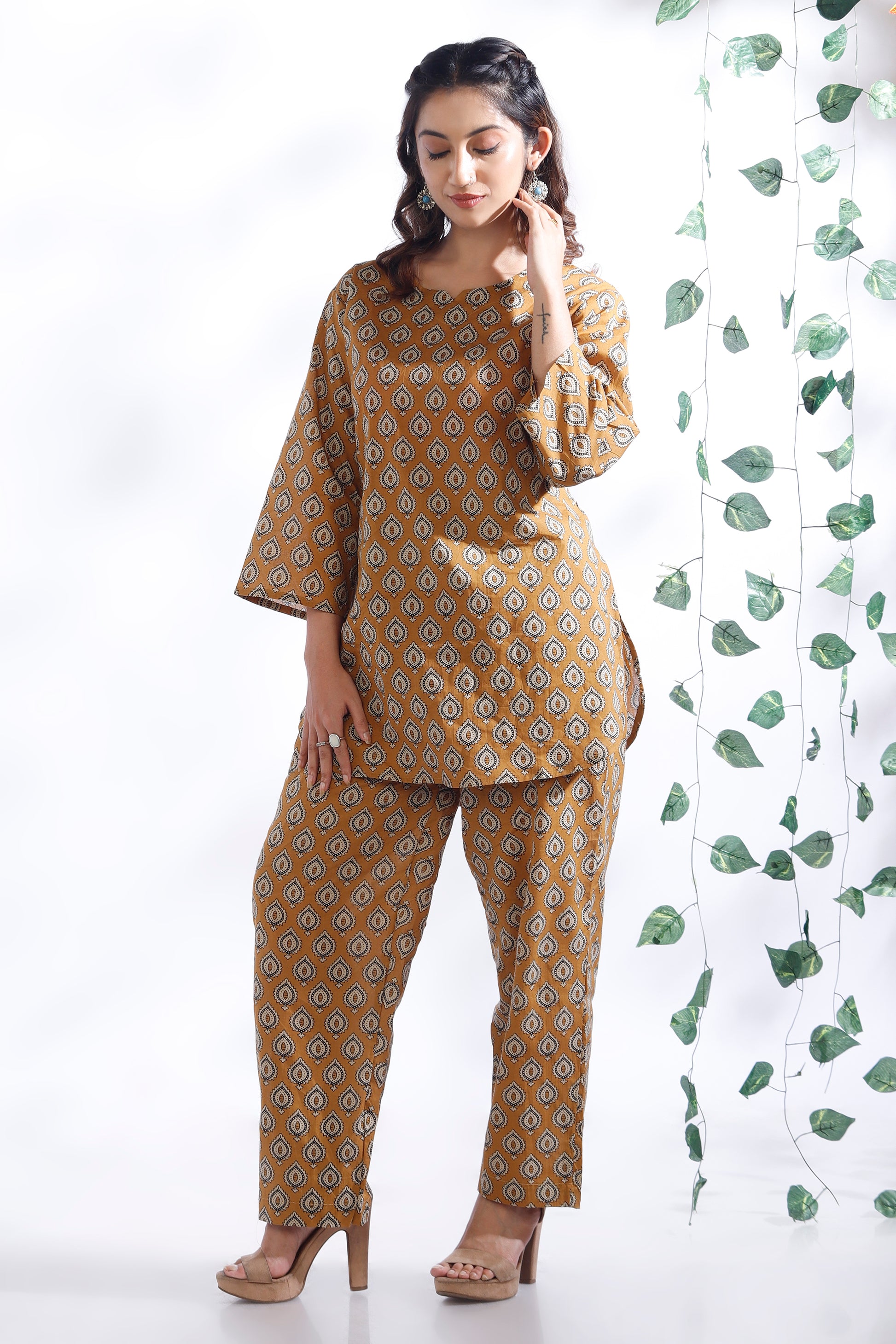 Woman wearing a mustard yellow patterned outfit with a white background and green leafy curtain.