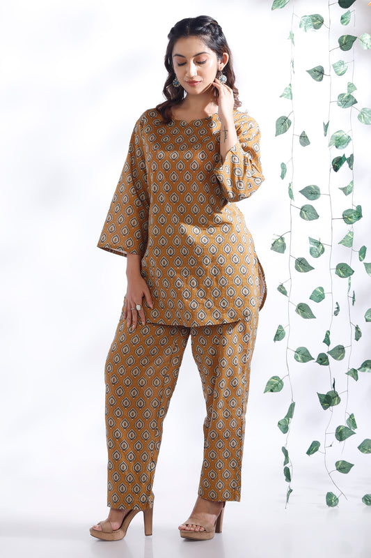 Woman wearing a mustard yellow patterned outfit with a white background and green leafy curtain.