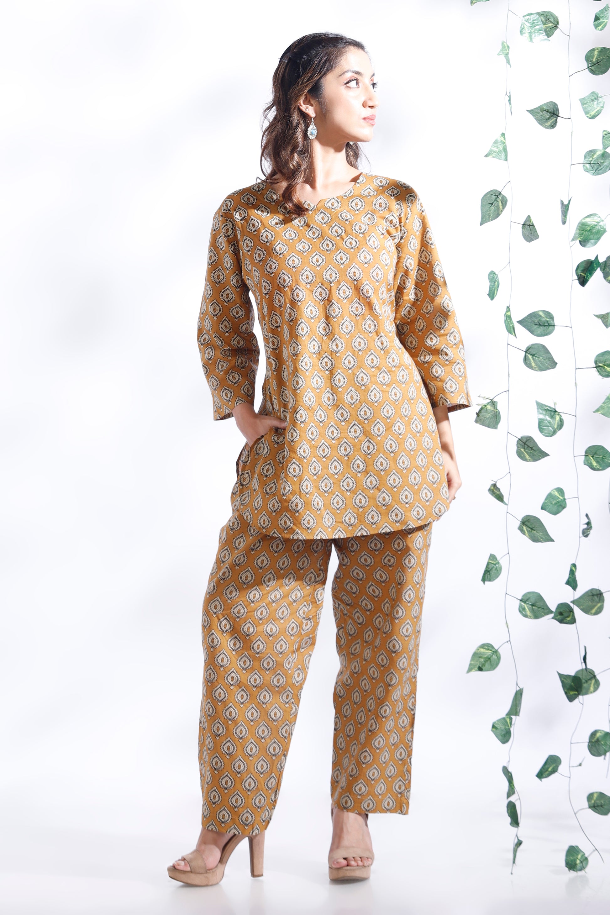 Woman wearing a yellow patterned outfit standing next to a white wall with green leaf designs.