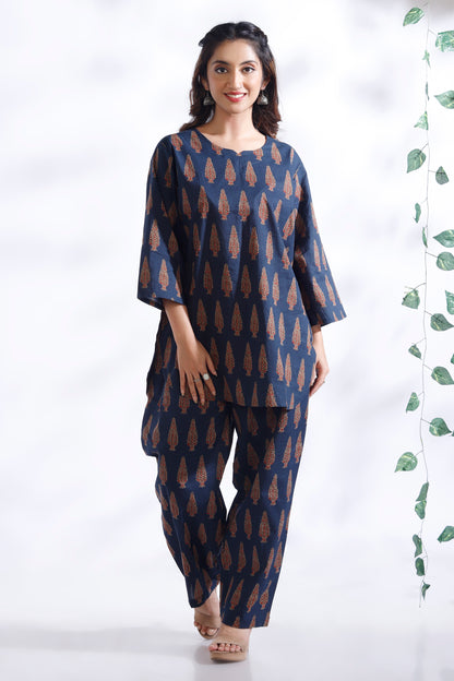 Woman wearing a blue patterned outfit standing against a white background with a plant on the side.
