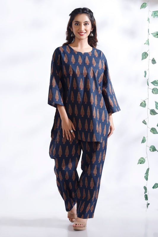 Woman wearing a blue patterned outfit standing against a white background with a plant on the side.