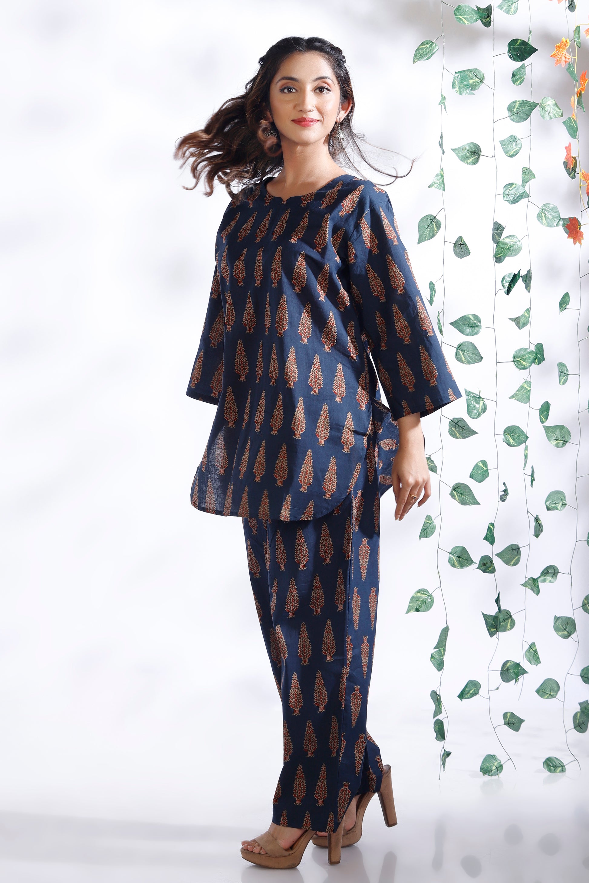 Woman wearing a blue and brown patterned outfit standing against a white background with a decorative plant.