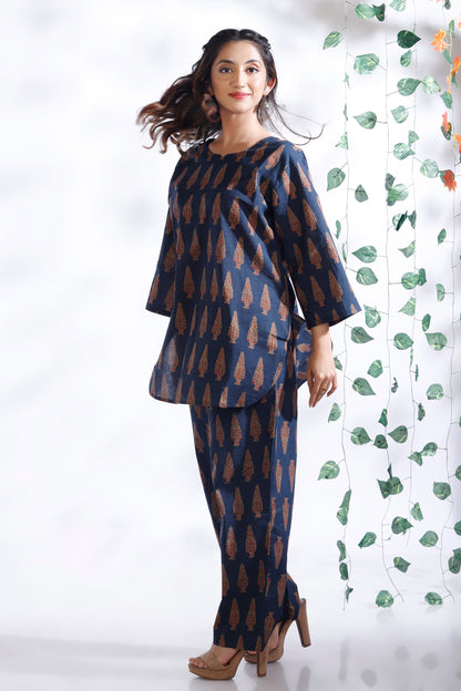 Woman wearing a blue and brown patterned outfit standing against a white background with a decorative plant.