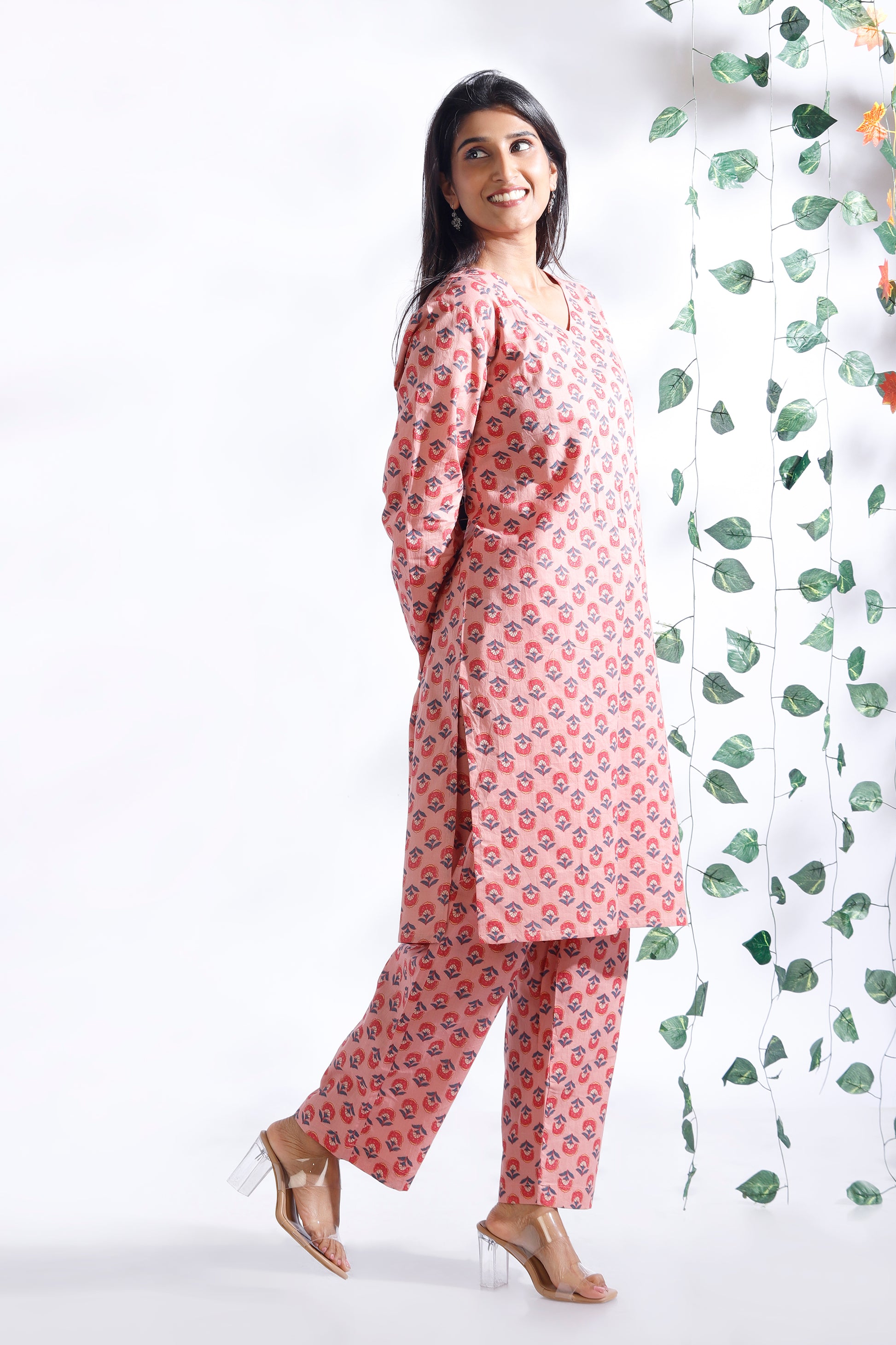 Woman wearing a pink floral outfit standing next to a decorative plant against a white background
