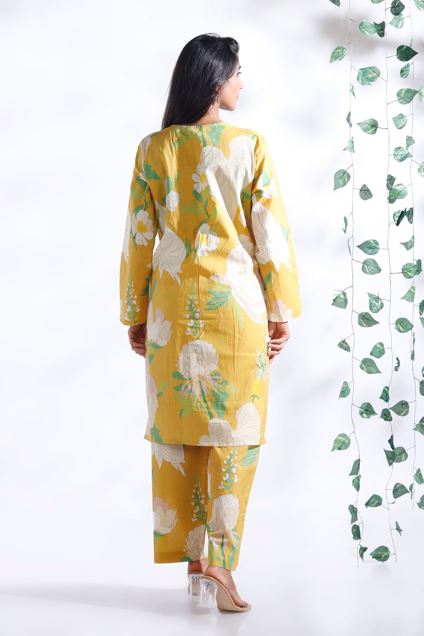 Woman wearing a yellow floral outfit standing next to a decorative plant against a white background