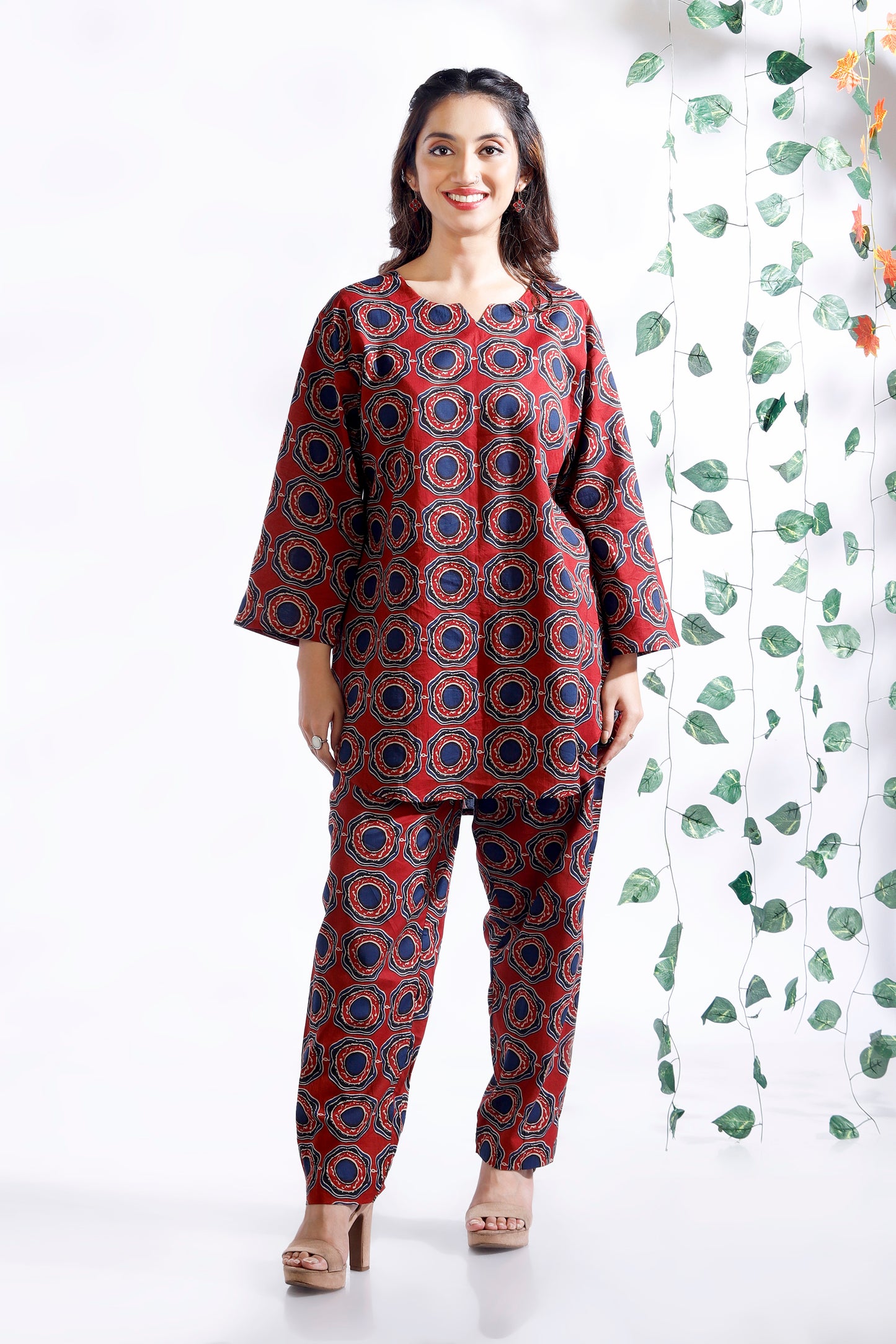 Woman wearing a red and blue patterned outfit standing against a white background with green leafy patterns.
