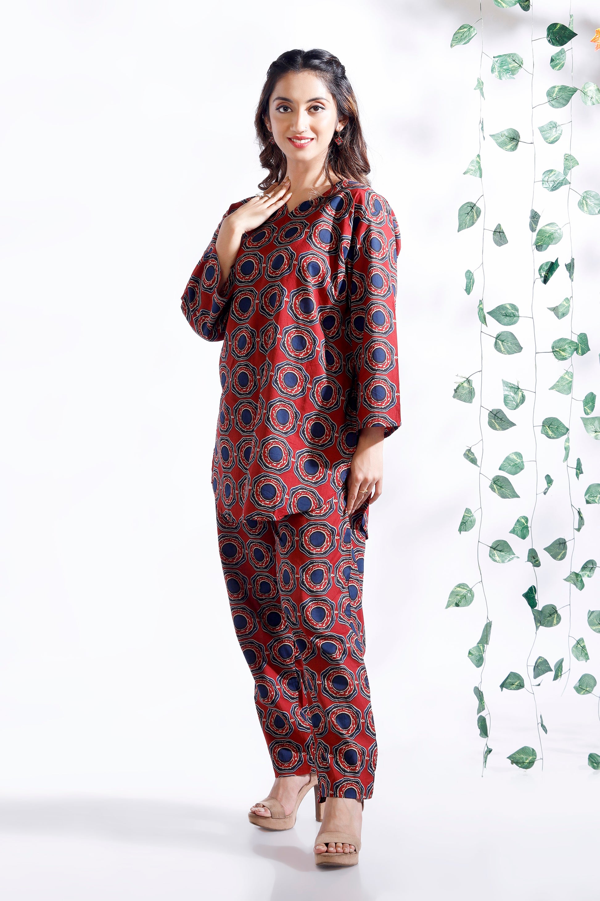 Woman wearing a red and blue patterned outfit standing next to a green leafy plant on a white background
