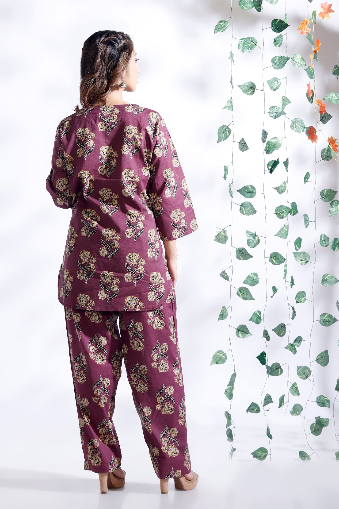 Woman wearing a purple floral outfit standing next to a white curtain with green leaf patterns.