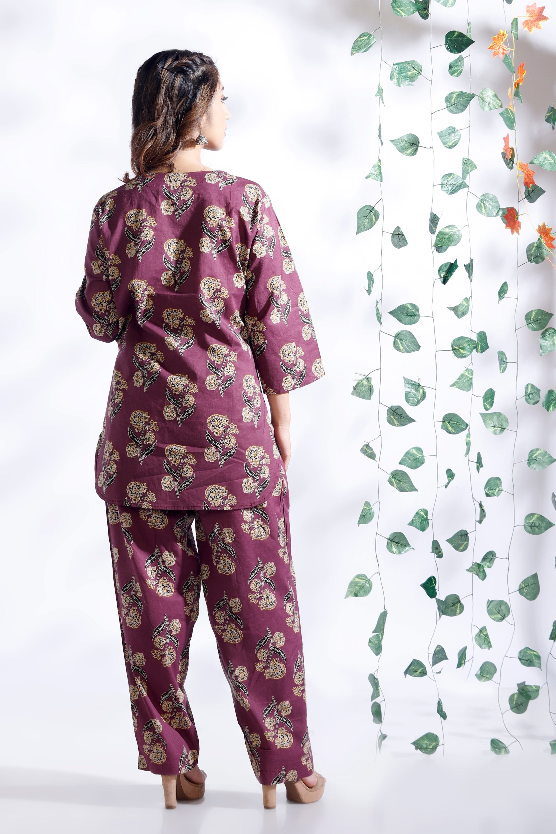 Woman wearing a purple floral outfit standing next to a white curtain with green leaf patterns.