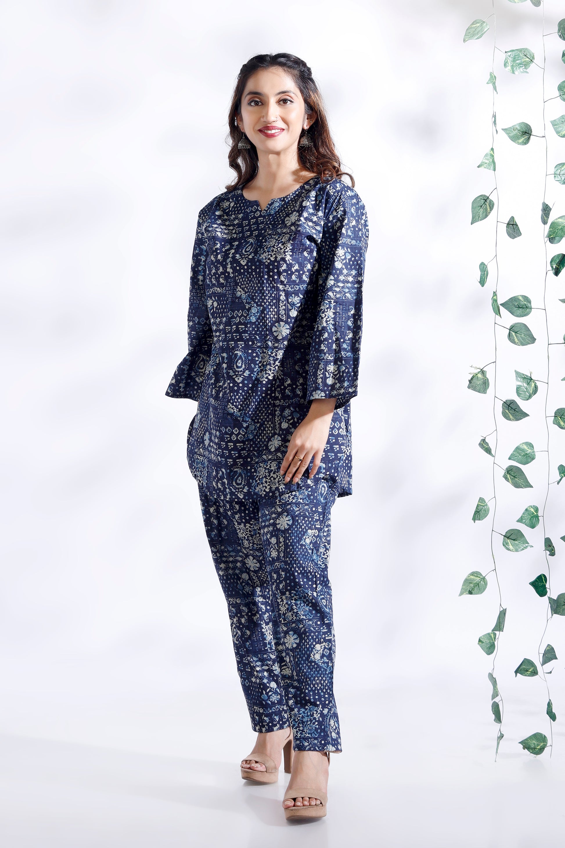 Woman wearing a blue patterned outfit standing against a white background with green leaves.