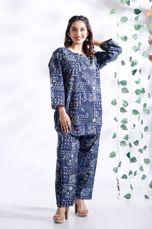 Woman wearing a blue patterned outfit standing next to a white curtain with green leaves.