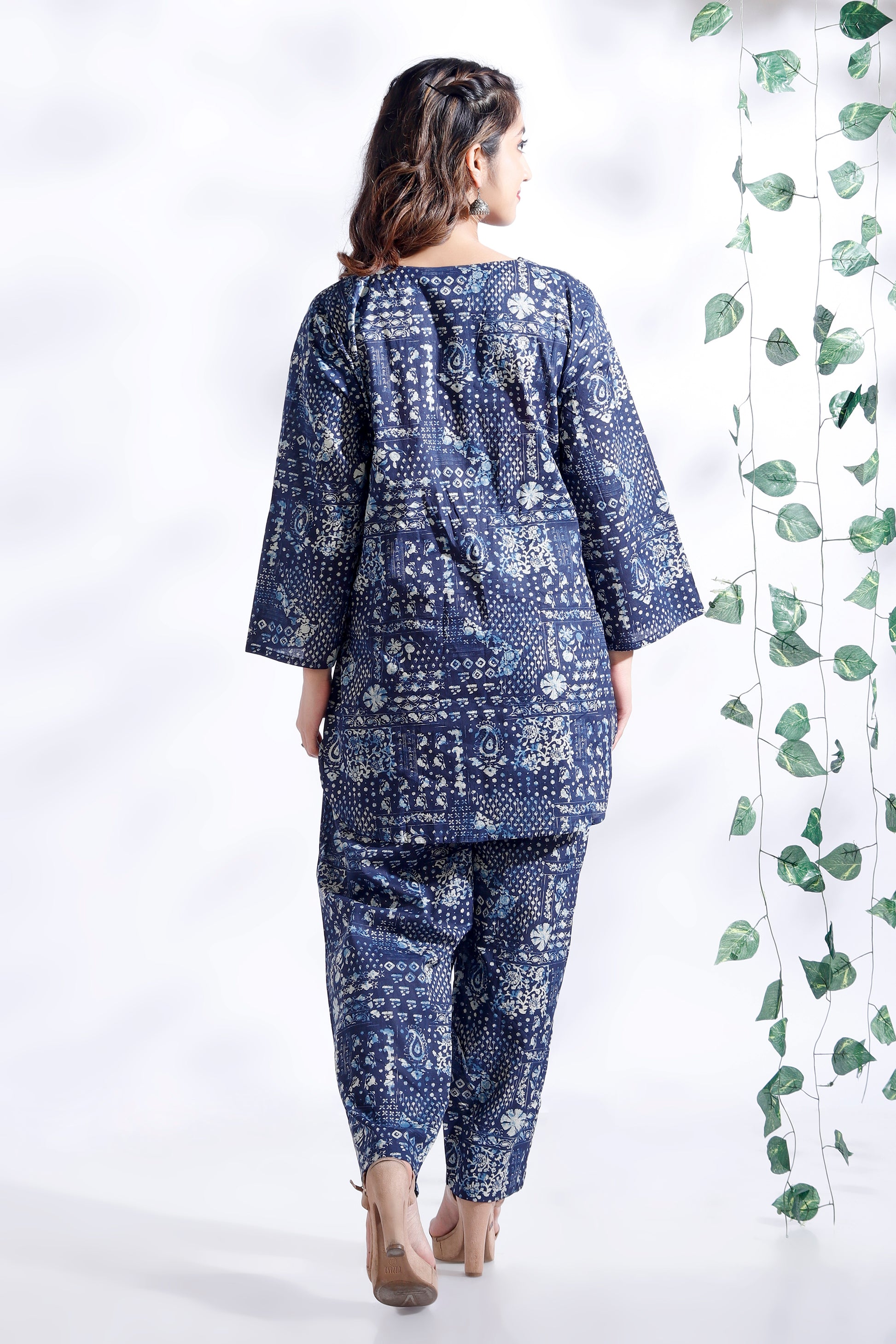 Woman wearing a blue patterned outfit standing against a white background with green leaves.