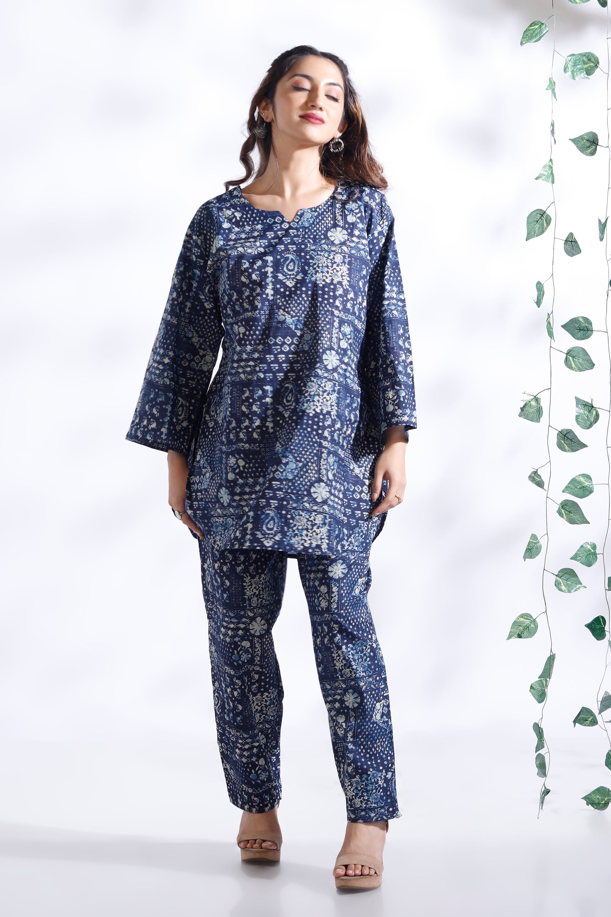 Woman wearing a blue patterned outfit standing against a white background with green leaves on the side.