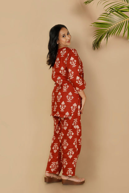 Woman wearing a red floral outfit standing against a beige wall with a plant.