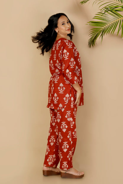 Woman wearing a red patterned outfit standing against a beige wall with a plant in the corner.