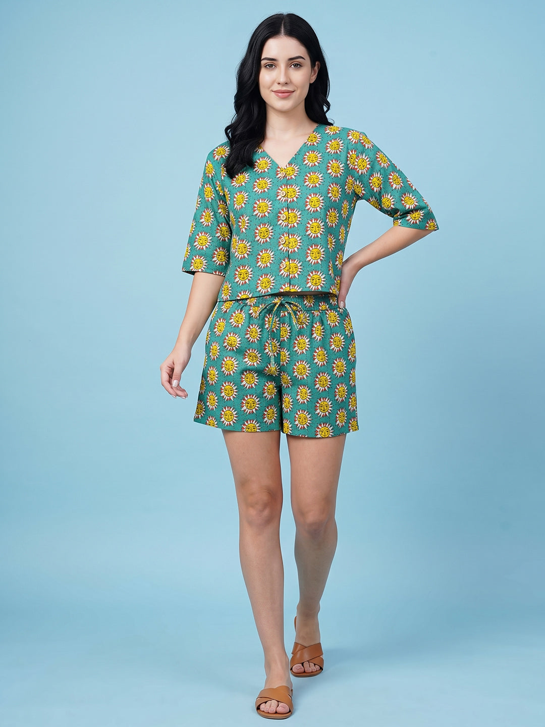 A woman modeling a matching two-piece set consisting of a short-sleeve V-neck top and coordinating shorts, both featuring a yellow sunflower print on a teal background, set against a solid light blue studio backdrop.