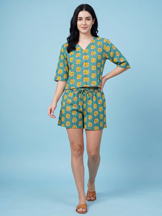 A woman modeling a matching two-piece set consisting of a short-sleeve V-neck top and coordinating shorts, both featuring a yellow sunflower print on a teal background, set against a solid light blue studio backdrop.