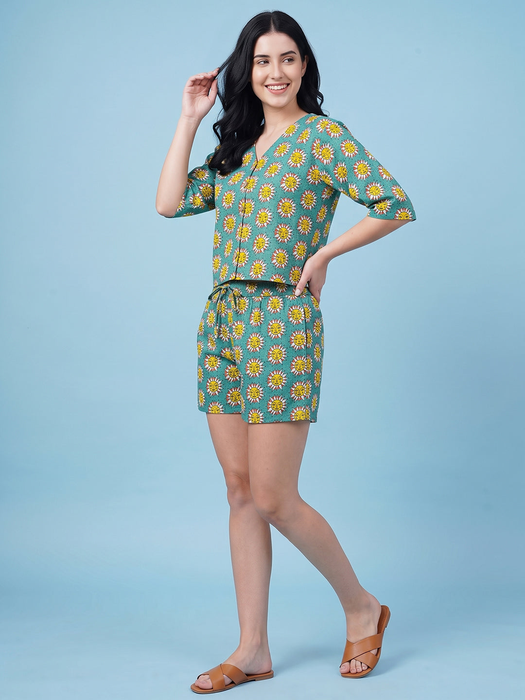 A woman modeling a matching two-piece set consisting of a short-sleeve V-neck top and coordinating shorts, both featuring a yellow sunflower print on a teal green background. She is posing against a solid light blue studio backdrop.