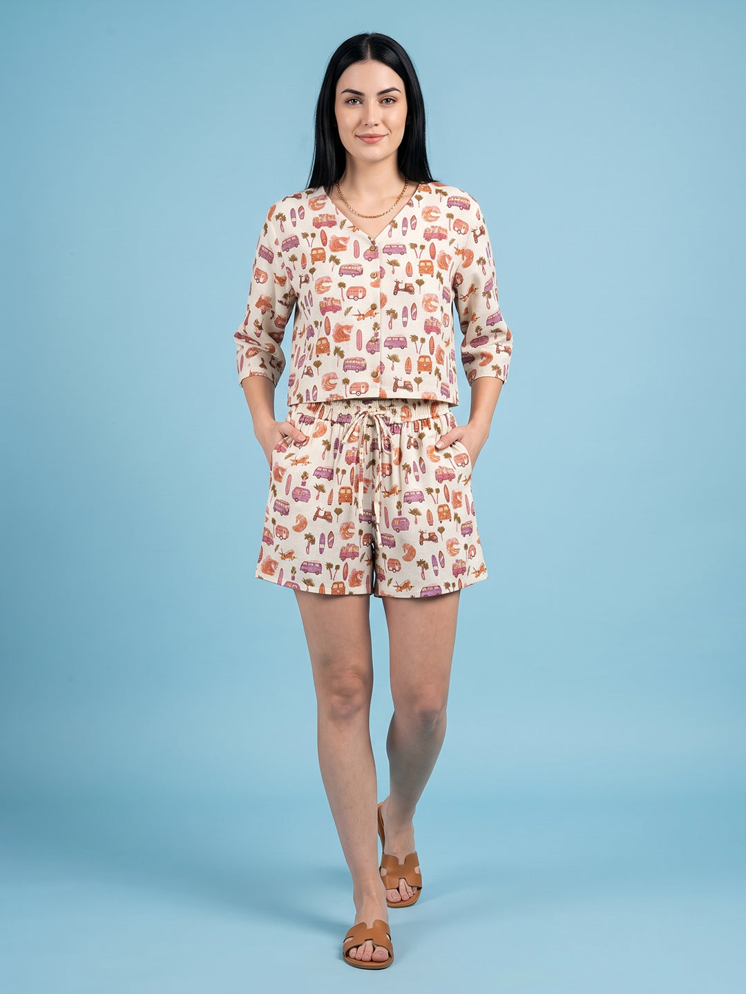 A woman modeling a matching two-piece lounge set featuring a long-sleeve top and shorts with an all-over whimsical print, set against a solid light blue studio background.