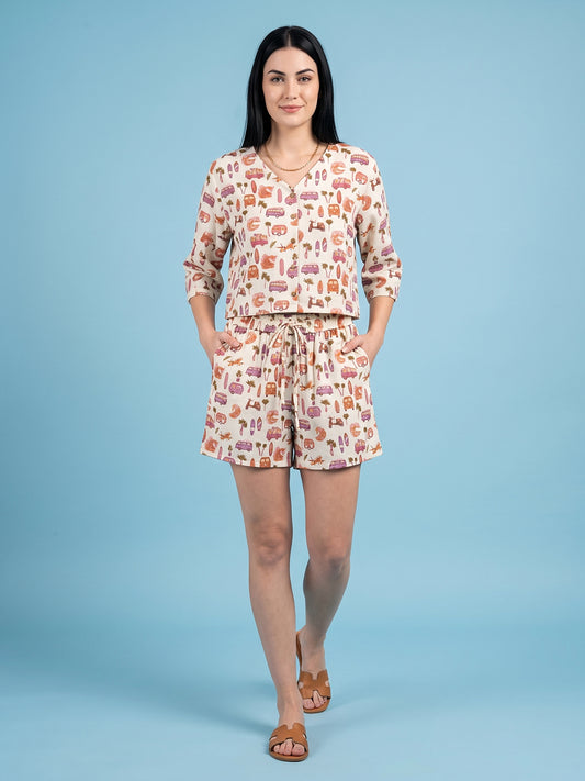 A woman modeling a matching two-piece lounge set featuring a long-sleeve top and shorts with an all-over whimsical print, set against a solid light blue studio background.