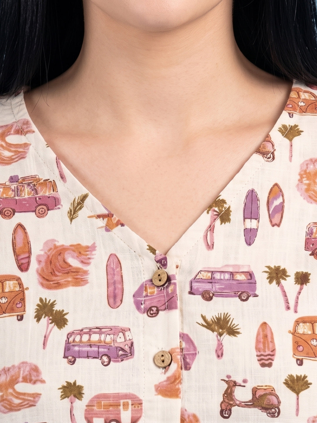 Close-up of a person wearing a cream-colored V-neck top with a retro pattern of purple vans, surfboards, palm trees, and waves. The top features a button placket with brown wooden buttons.