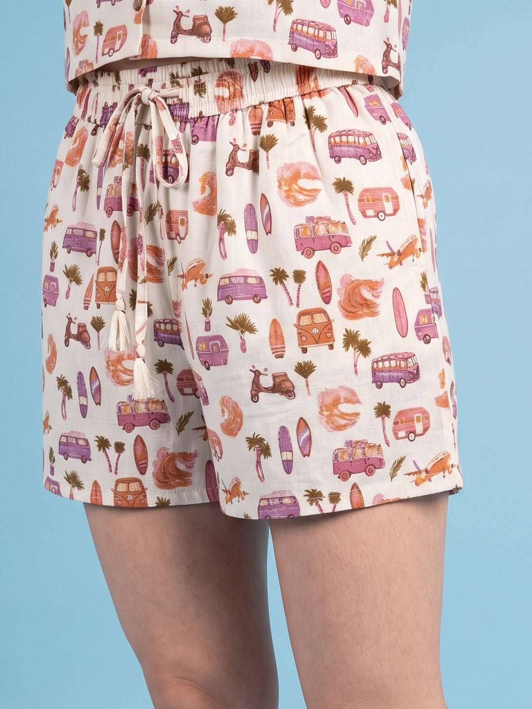 A person wearing high-waisted shorts with an all-over retro beach-themed print featuring purple campers, orange surfboards, pink scooters, and brown palm trees on a cream background, paired with a matching top against a solid light blue studio background.