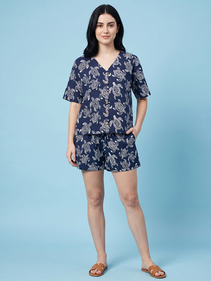 A woman modeling a navy blue two-piece loungewear set with a white floral print, consisting of a short-sleeve V-neck top and matching shorts, paired with brown slide sandals against a solid light blue background.