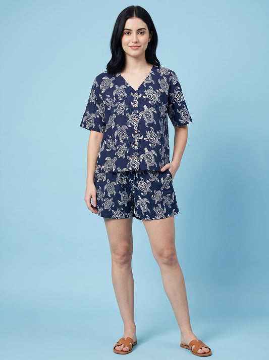 A woman modeling a navy blue two-piece loungewear set with a white floral print, consisting of a short-sleeve V-neck top and matching shorts, paired with brown slide sandals against a solid light blue background.