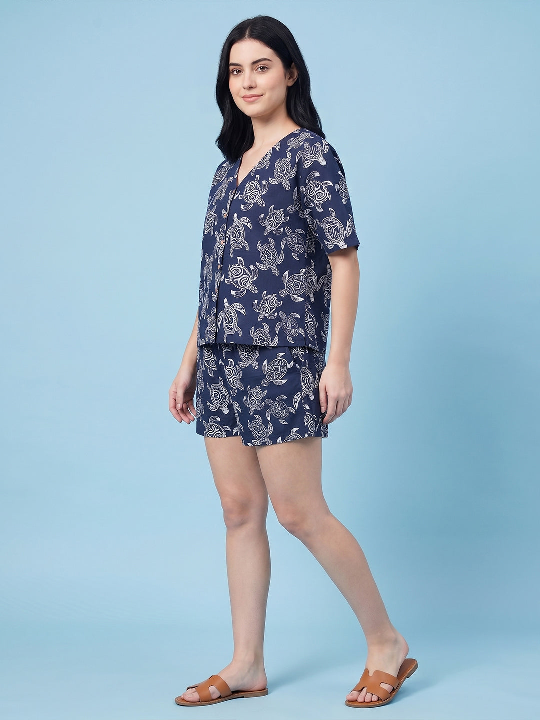 A woman modeling a navy blue two-piece pajama set with a white floral print, consisting of a short-sleeve top and matching shorts, paired with brown slide sandals against a solid light blue background.