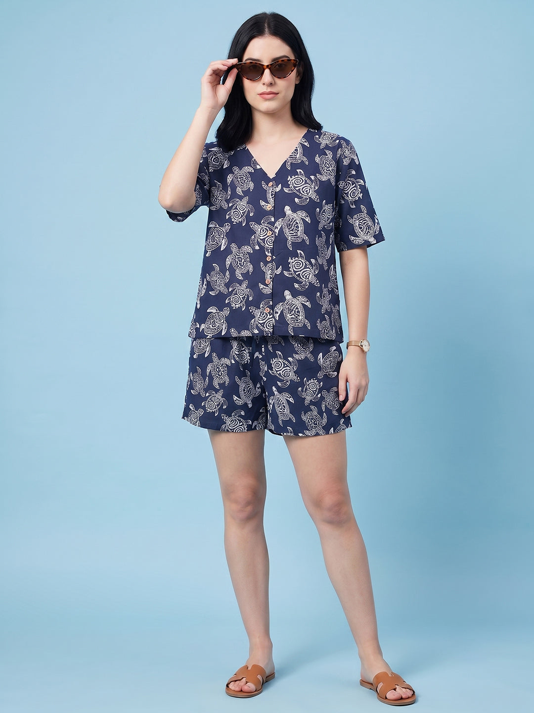 A woman modeling a navy blue two-piece lounge set with a white abstract pattern, consisting of a short-sleeve V-neck top and matching shorts, styled with brown sunglasses and tan slide sandals against a solid light blue background.