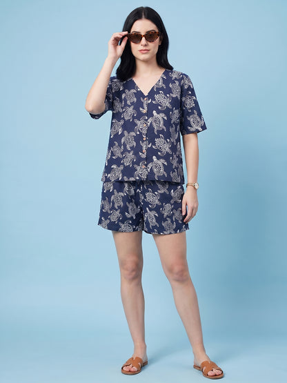 A woman modeling a navy blue two-piece lounge set with a white abstract pattern, consisting of a short-sleeve V-neck top and matching shorts, styled with brown sunglasses and tan slide sandals against a solid light blue background.